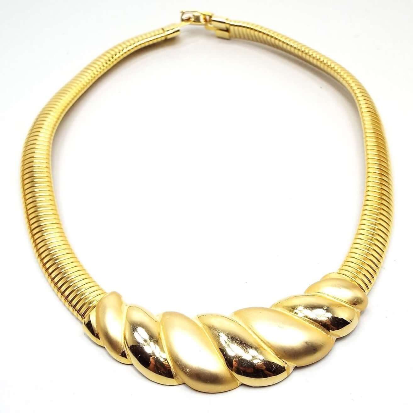 Front view of the retro vintage bib choker necklace. Metal is gold tone in color. Bottom bib part has an angled scalloped style design with alternating matte and shiny metal. Each side has a wide omega chain that has curved linked bars going all the way up to the snap lock clasp.