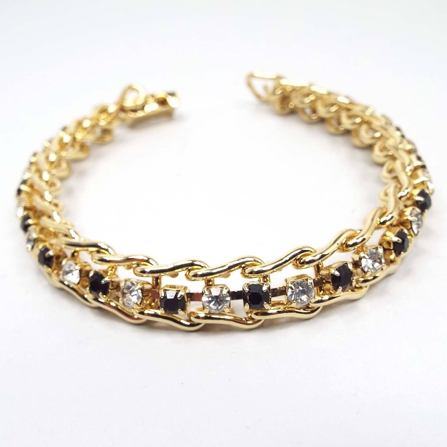 Front view of the retro vintage rhinestone bracelet. The metal is shiny gold tone in color and has sideways curved U shaped links. Inside each link is a small round rhinestone. The rhinestones alternate in color down the length of the bracelet in black and sparkling clear. There is a snap lock clasp at the end.