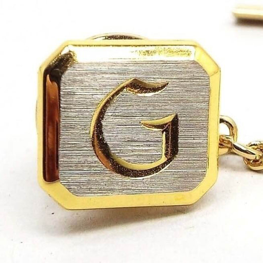 Front view of the two tone Mid Century vintage initial tie tack. The front of the tie tack is matte silver tone color with a gold tone color block letter initial G engraved on the front. The rest of the tie tack is gold tone in color. There is a chain coming off the back clutch that has a small bar on the end.
