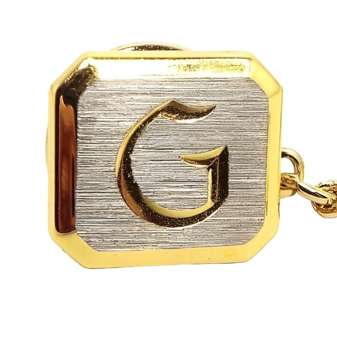Front view of the two tone Mid Century vintage initial tie tack. The front of the tie tack is matte silver tone color with a gold tone color block letter initial G engraved on the front. The rest of the tie tack is gold tone in color. There is a chain coming off the back clutch that has a small bar on the end.