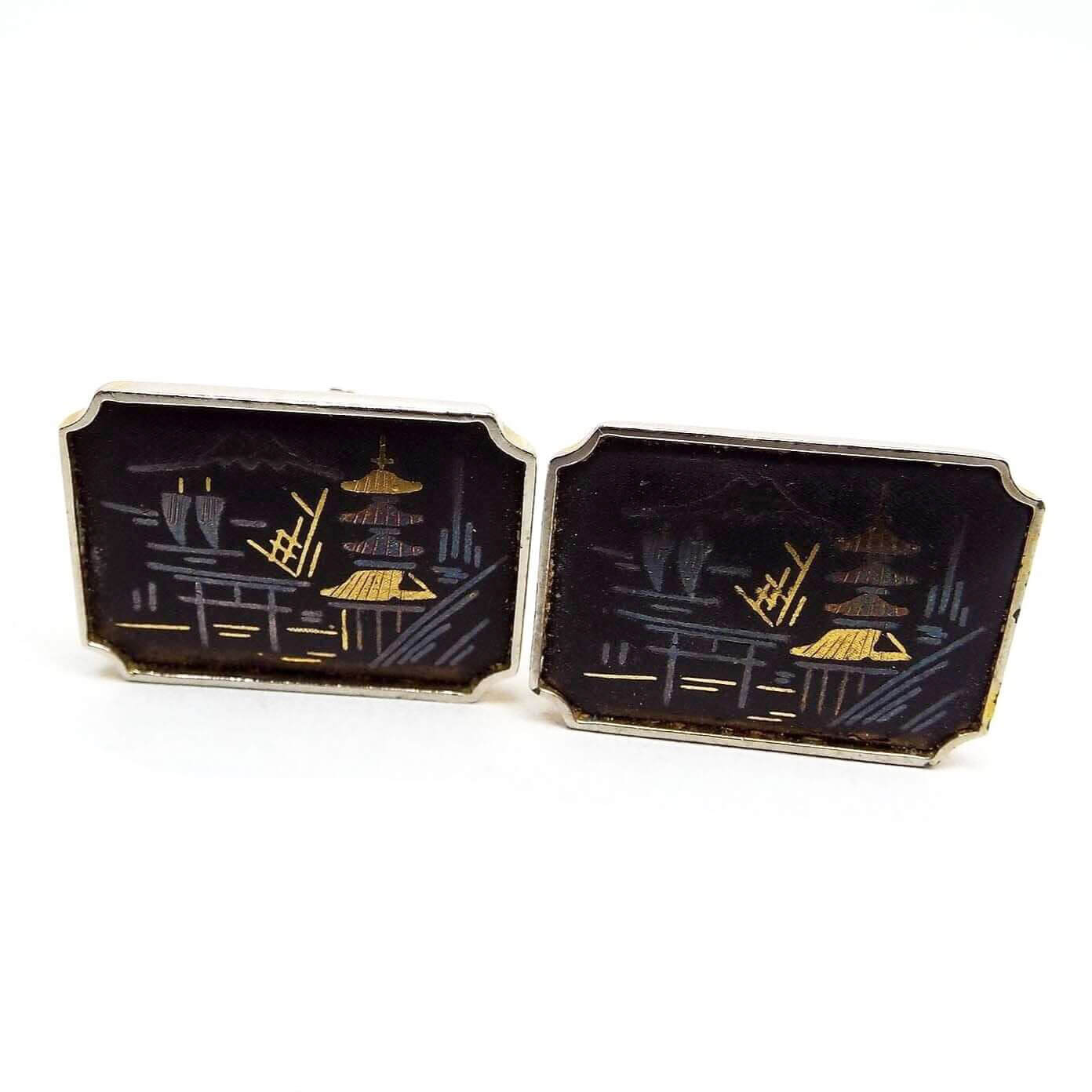Front view of the Amita Damascene cufflinks. Front are black painted with etched design of a Japanese home on the water with a boat in the background. The engraved area shows metal beneath in gold and silver color making up the sea side design. They are rectangular in shape with curved indented corners. 
