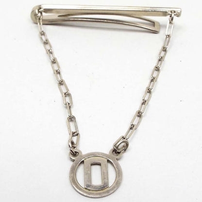 Front view of the Mid Century vintage tie bar chain. The metal is silver tone in color. The top bar has a very thin line design on the front and a curved back bar with open middle. The back bar is cuved outwards some, so it would be better for thicker style ties. Small cable chain with rectangle links comes down from each side of the bar and there is a round open charm at the bottom with a block letter initial O in the middle.