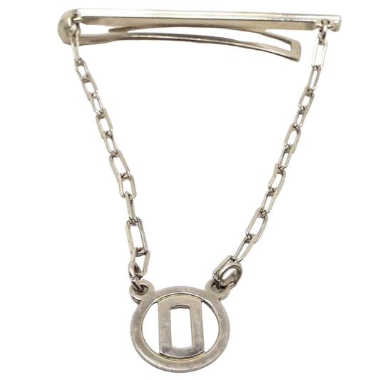 Front view of the Mid Century vintage tie bar chain. The metal is silver tone in color. The top bar has a very thin line design on the front and a curved back bar with open middle. The back bar is cuved outwards some, so it would be better for thicker style ties. Small cable chain with rectangle links comes down from each side of the bar and there is a round open charm at the bottom with a block letter initial O in the middle.