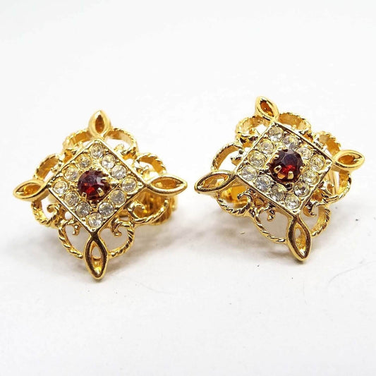 Front view or the retro vintage Avon clip on earrings. They are diamond shaped with a filigree open fancy scroll like design around the outer edge. There is an inner diamond shaped area with pavé set small round clear rhinestones. There is a larger sized round deep red rhinestone in the middle. 