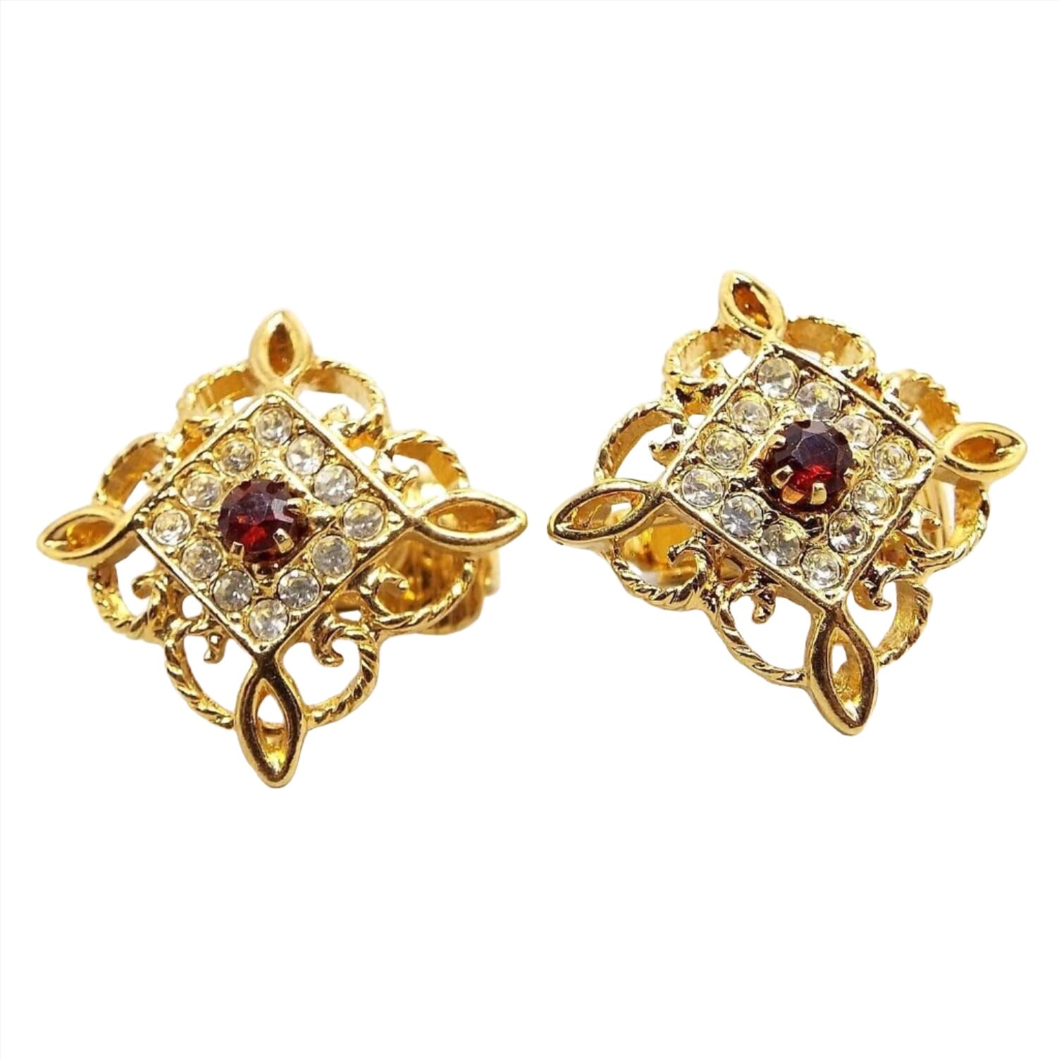 Front view or the retro vintage Avon clip on earrings. They are diamond shaped with a filigree open fancy scroll like design around the outer edge. There is an inner diamond shaped area with pavé set small round clear rhinestones. There is a larger sized round deep red rhinestone in the middle. 