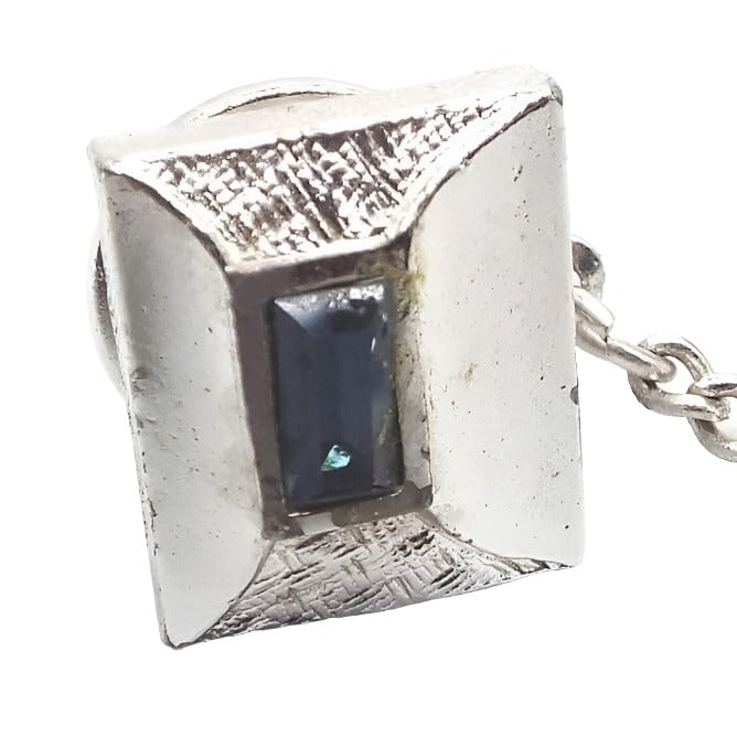 Front view of the Mid Century vintage rhinestone tie tack. The metal is silver tone in color. it has a rectangle design that is sloped outward off center and slightly on the left. The middle of the slope has a single baguette rhinestone in dark blue. The top and bottom of the slope are textured and the sides are smooth. The clutch on the back has a chain with a bar at the end.
