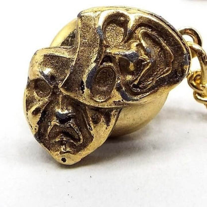 Front view of the Mid Century vintage comedy and tragedy mask tie tack. It is dark gold tone in color and has two raised 3D style mask type faces angled towards one another. One is happy and smiling and the other is frowning. The back clutch has a chain with a small bar at the end.