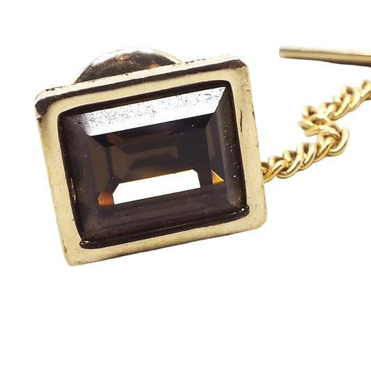 Enlarged front view of the Mid Century vintage Swank rhinestone tie clip. It's shaped like a rectangle and is gold tone in color. There is a larger sized faceted brown rectangle rhinestone in the middle. A tiny scratch in the gold tone metal can be see on the right side front of the bezel area when magnified.
