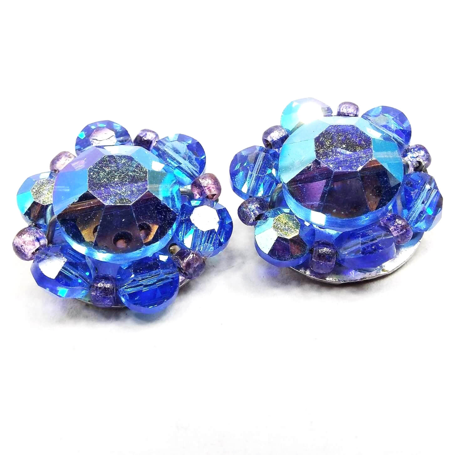 Front view of the Mid Century vintage AB blue crystal glass beaded clip on earrings. They are blue with hints of other colors from the aurora borealis coating. There are large faceted flat back rhinestone shaped beads in the middle surrounded by smaller ones and seed beads in a slightly different shade of blue.