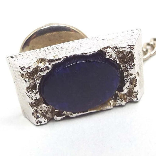 Front view of the Mid Century vintage sodalite tie tack. The dark blue sodalite gemstone cab is oval in shape and is surrounded by a nugget style textured silver tone rectangle setting. The clutch on the back has a chain with a small bar on the end.