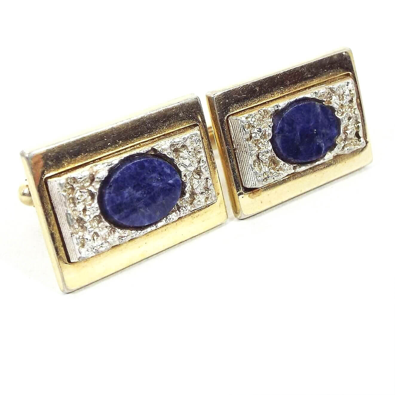 Angled front view of the retro vintage Anson sodalite cufflinks. The main color of the setting is gold tone. They are rectangular shaped with bumpy textured silver tone rectangles on the front. There are oval denim blue marbled sodalite gemstone cabs in the very middle. 
