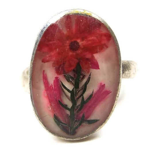 Picture of front of the ring. Ring top is oval with silver color edge. There is a flower in the middle. Top of flower has lots of petals with orange, pink, and red hues. Stem of flower is green with spikey style leaves coming off of it that are angled up. At the bottom there are additional petals by themselves that are spiked upwards in pink hues. The entire middle is encased in clear resin. 