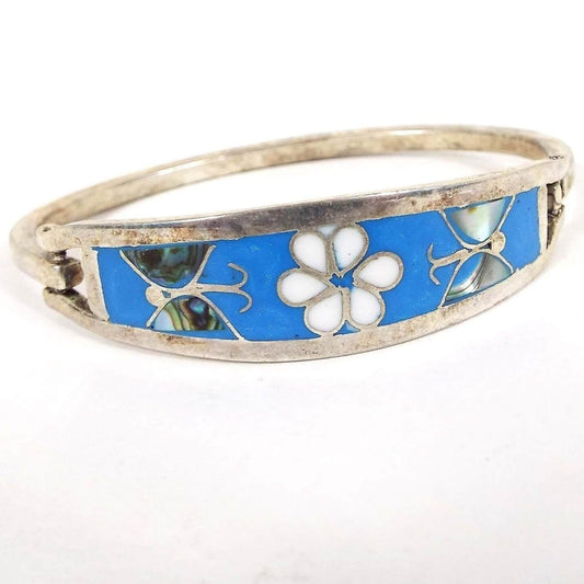 Angled front and top view of the retro vintage Taxco hinged bangle bracelet. The metal is silver tone in color. There is a large curved area on top with bright blue enamel. In the middle is a white enameled flower that has a butterfly design on each side. The butterfly wings have inlaid pearly multi color abalone shell. The back of the bracelet is a squared bar that curves around and is hinged on one side and has a hook on the other to open and close the bracelet.