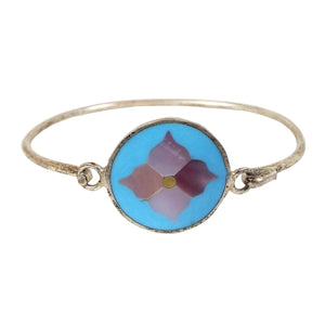 Hinged bangle bracelet is silver in color with a thin rounded band of metal that goes in a circle around the back. On the front is a round disc that has light blue resin with pieces of inlaid mother of pearl in the middle to form a flower shape. Flower has shades of pearly pink. One side of the disc has the metal band permanently curved onto a loop and the other side has just a curve where you can hook or unhook it from the loop to put the bracelet on or take it off.