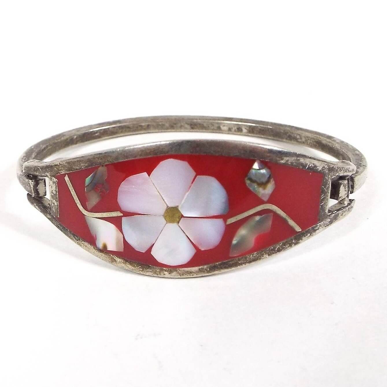 Front view of the Mexican Alpaca hinged bangle has two bands of metal and a large area in the middle with tapered ends. It has red resin with a flower design in the middle.Flower petals are mother of pearl shell and pearly white in color. Leaves on each side of the flower are abalone shell and are pearly multi color. There is a thin band of metal curved around the back and one side has a hook that clicks onto one side of the bangle to take it on and off. Metal is a darkened silver color from age.