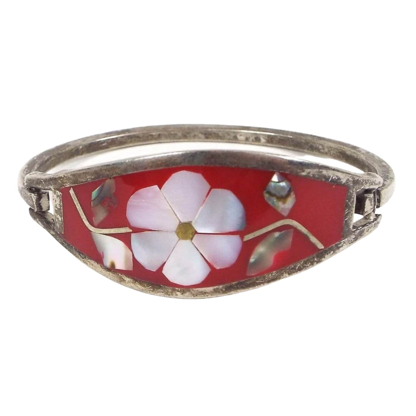 Front view of the Mexican Alpaca hinged bangle has two bands of metal and a large area in the middle with tapered ends. It has red resin with a flower design in the middle.Flower petals are mother of pearl shell and pearly white in color. Leaves on each side of the flower are abalone shell and are pearly multi color. There is a thin band of metal curved around the back and one side has a hook that clicks onto one side of the bangle to take it on and off. Metal is a darkened silver color from age.