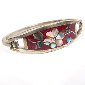 Angled front and side view of the retro vintage Mexican hinged bangle bracelet. The front has a large curved area with pearly pink and red marbled resin enamel. The middle has a flower design with pieces of multi color pearly inlaid abalone shell making up the petals and leaves on either side. The metal is silver tone in color and has a curved band that is hinged to one side of the top piece and has a slight hook on the other to open and close the bracelet.