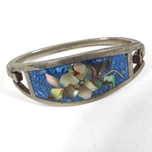 Angled front and side view of the retro vintage Mexican hinged bangle bracelet. The front has a large curved area with resin and tiny blue dyed stone chips. The middle has a flower design with pieces of multi color pearly inlaid abalone shell making up the petals and leaves on either side. The metal is silver tone in color and has a curved band that is hinged to one side of the top piece and has a slight hook on the other to open and close the bracelet.