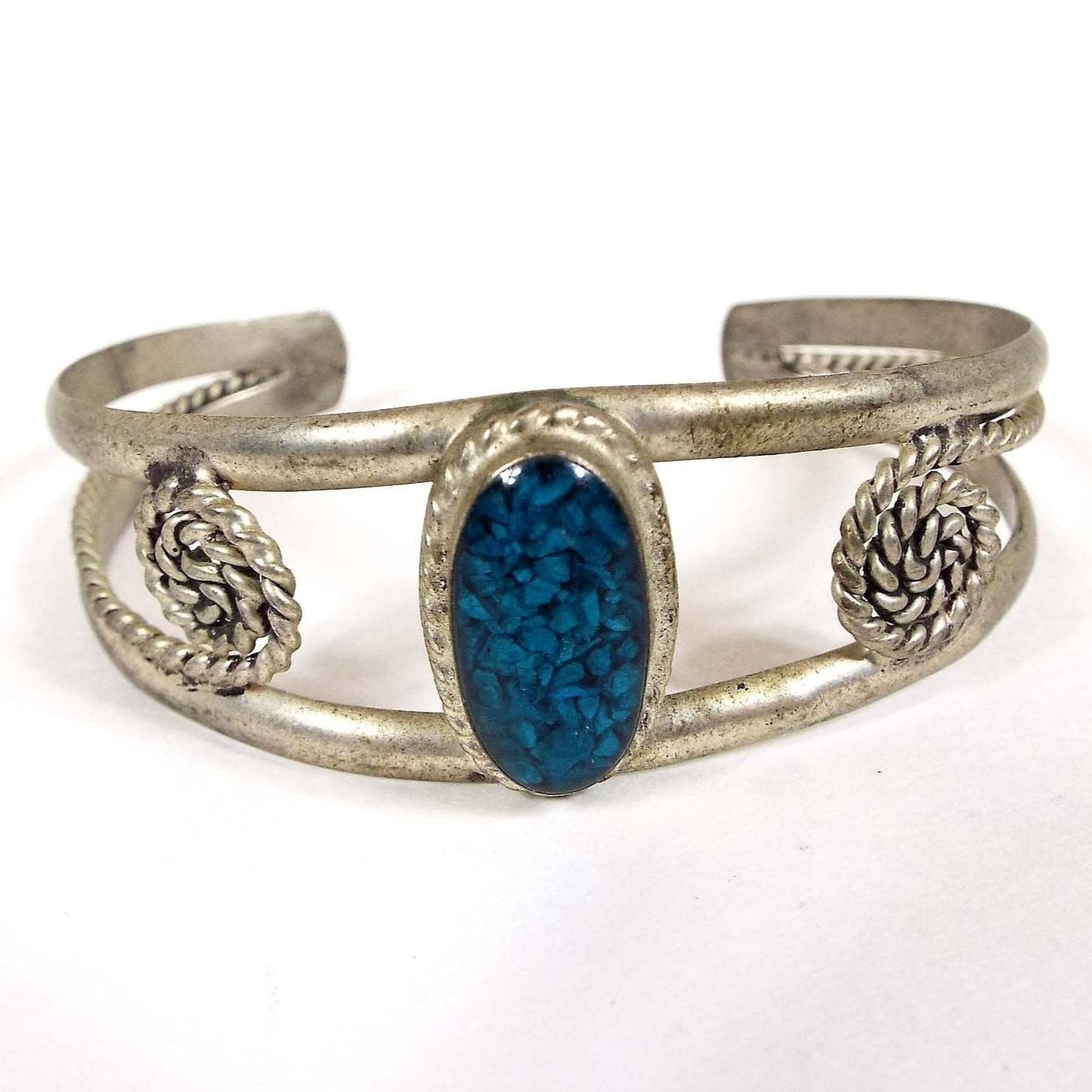 Front view of the retro vintage Mexican alpaca cuff bracelet. Metal is silver tone in color. The top middle has an oval bezel with blue resin and small blue gemstone chips. There is a band of metal at the top and bottom of the oval that curves around to the back to form the cuff. It has tapered ends and from the ends is a smaller twisted wire that comes back towards the front and then spirals curling inwards.