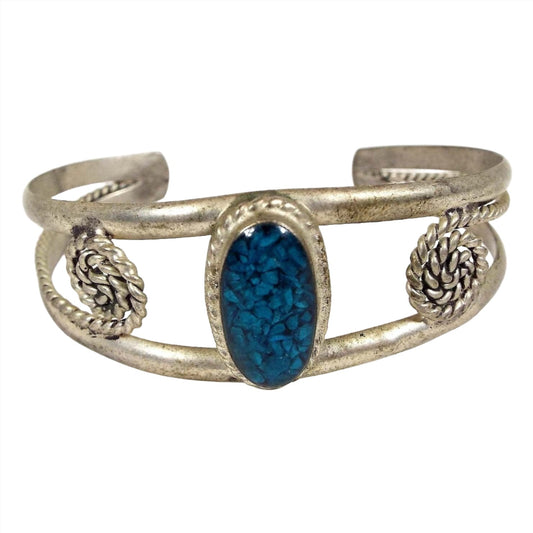 Front view of the retro vintage Mexican alpaca cuff bracelet. Metal is silver tone in color. The top middle has an oval bezel with blue resin and small blue gemstone chips. There is a band of metal at the top and bottom of the oval that curves around to the back to form the cuff. It has tapered ends and from the ends is a smaller twisted wire that comes back towards the front and then spirals curling inwards.