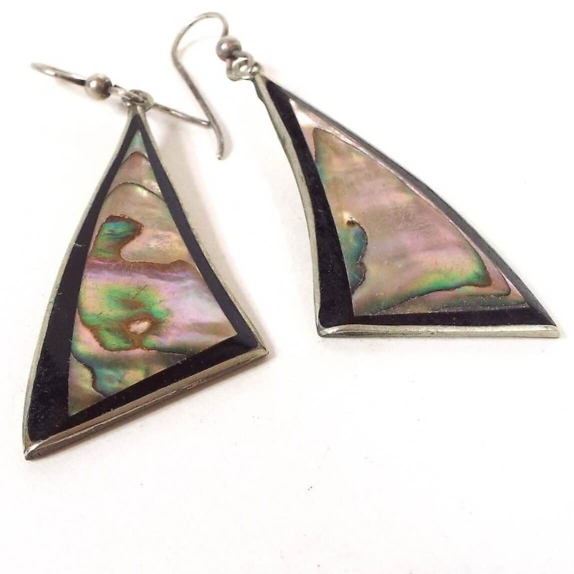Front side of the Mexican Alpaca earrings. The metal is a darker silver in color from age. The drops are curved triangles with large pieces of pearly multi color inlaid abalone shell. The edge around the abalone is black resin enamel. Tops have pierced fish hook style ear wires.