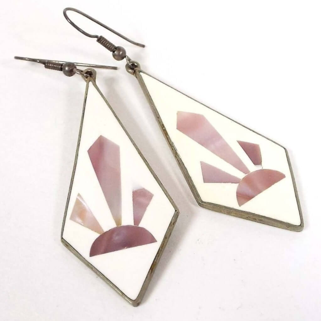 Front view of the Alpaca marked earrings. Earring drops are angled teardrop shaped with white enamel. Each one has a sun ray or cactus type Southwestern style design in pearly pink color mother of pearl shell. Tops have curved fish hook style ear wires. The metal is darkened silver in color.