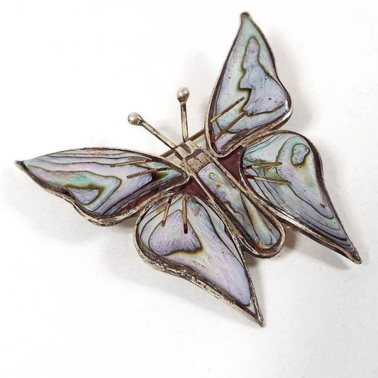 Front view of the Mid Century vintage Taxco butterfly brooch pin. The metal is silver tone in color. The body of the butterfly has inlaid multi color abalone shell. It has a coating to give it a mostly matte pastel like appearance.