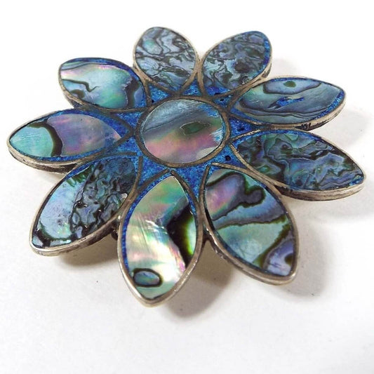 Front view of the retro vintage Taxco sterling silver brooch pendant. The sterling is slightly darkened from age. The brooch has a floral design that has pearly multi color inlaid abalone shell for the petals and middle part of the flower. In between the petals and middle is a small area of tiny inlaid blue dyed stone chips.