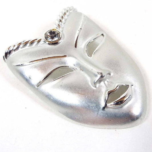 Front view of the retro vintage mask brooch pin. The metal is mostly matte silver tone in color with shiny silver tone color accents around the open cut out facial features. The top part angle's inward like a V shape and has a single round clear rhinestone in the middle. 