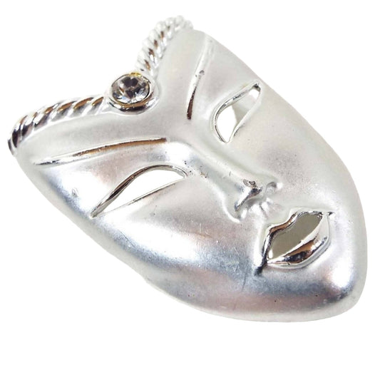 Front view of the retro vintage mask brooch pin. The metal is mostly matte silver tone in color with shiny silver tone color accents around the open cut out facial features. The top part angle's inward like a V shape and has a single round clear rhinestone in the middle. 
