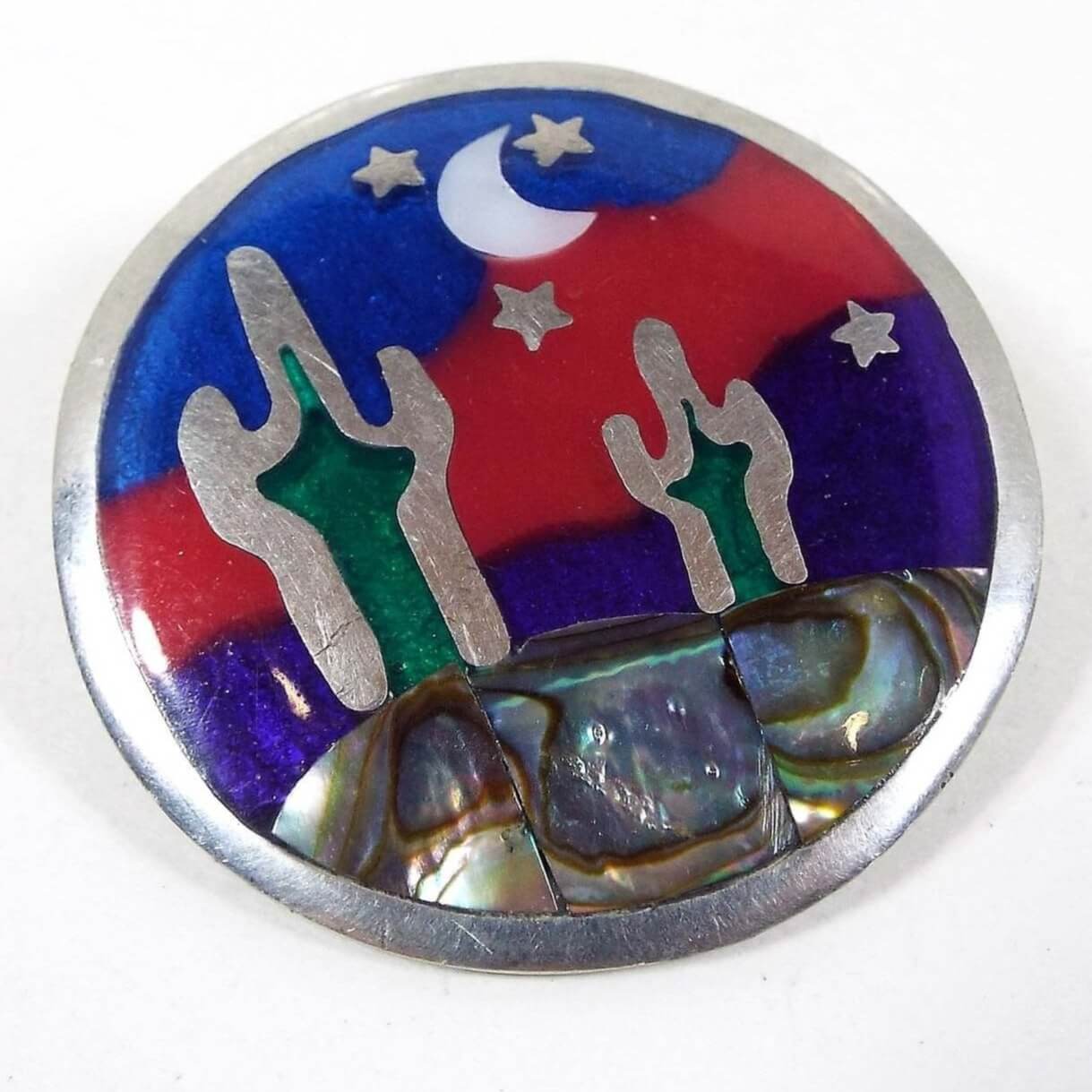 Front view of the retro vintage Southwestern style desert brooch pendant. The metal is silver tone in color. The brooch is round with a night time desert scene on the front. The top has tiny metal stars and an inlaid mother of pearl shell cab in the shape of a half moon. The bottom part of the brooch has pearly multi color inlaid abalone shell. There are two cactus with green enamel and the background has blue enamel with a wavy red enamel stripe through it.
