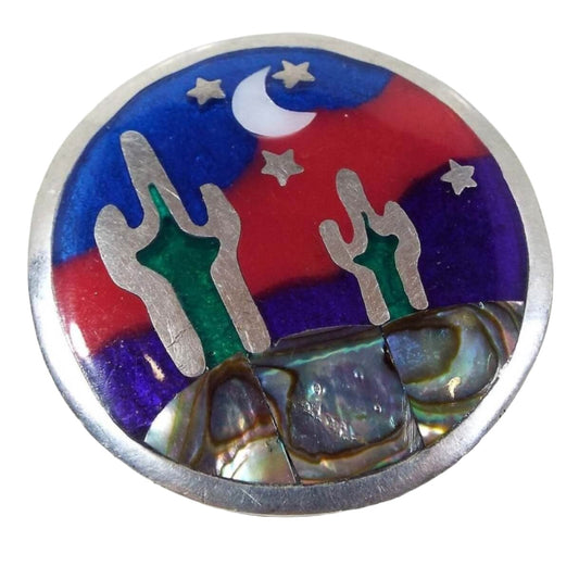 Front view of the retro vintage Southwestern style desert brooch pendant. The metal is silver tone in color. The brooch is round with a night time desert scene on the front. The top has tiny metal stars and an inlaid mother of pearl shell cab in the shape of a half moon. The bottom part of the brooch has pearly multi color inlaid abalone shell. There are two cactus with green enamel and the background has blue enamel with a wavy red enamel stripe through it.