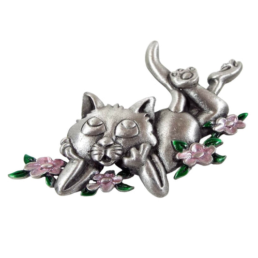 Front view of the retro vintage JJ brooch. It has a 3D raised style design of a cat laying on its stomach with its legs up in the air and its paws supporting its head as if it's daydreaming. The cat is surrounded with four flowers. The flowers are light pink enameled with green enameled leaves. The rest of the brooch is matte pewter gray metal.