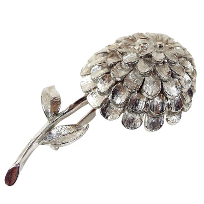 Front view of the 1960's Mid Century vintage floral brooch pin. The metal is textured matte silver tone in color. It is shaped like a dahlia flower with lots of petals on the head of the flower. The stem comes down from the bottom with a small leaf on each side.