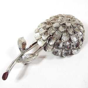 Front view of the 1960's Mid Century vintage floral brooch pin. The metal is textured matte silver tone in color. It is shaped like a dahlia flower with lots of petals on the head of the flower. The stem comes down from the bottom with a small leaf on each side.