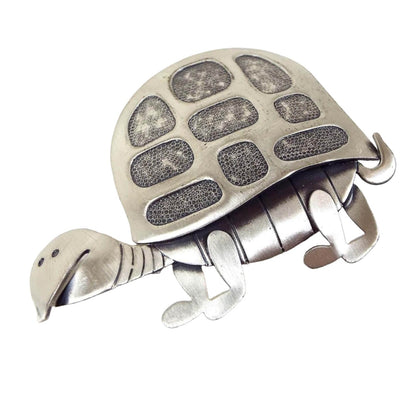 Front view of the Mid Century vintage Beau sterling silver tortoise brooch. The sterling is matte silver in color. The tortoise has flatter styled metal pieces comprising his body, legs, tail, and shell that are affixed together. The shell has oval and rectangle areas that are darker and have a small chain mesh like design on them. 