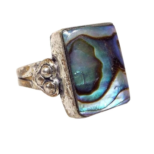 Ring is silver in color. It has a large rectangle on top with an abalone shell cab. The abalone has swirls of color in blue, green, and purple. The side of the ring has a heart shape with 3 round silver colored balls in it. The silver color on the ring is darkened , especially on the bottom part where the band is.