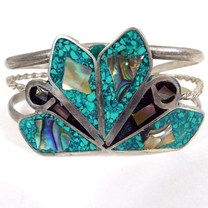 Front view of the retro vintage cuff bracelet. It is a wider silver tone bracelet with three curved bands to form the cuff. The top and bottom are rounded and the middle is thinner twisted wire. On the very top of the bracelet is a large area with angled teardops with small ends pointing inwards. Four of the larger teardrop shapes have tiny inlaid turquoise chips and a piece of inlaid abalone shell. The smaller ones have a curve of metal, black resin, and two small pieces of abalone shell. 