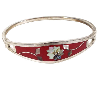 Front view of the retro vintage Taxco floral hinged bangle bracelet. The metal is silver tone in color. The top part has a long curved area with red enamel and a flower design in the middle. The flower petals and leaves are pearly multi color inlaid abalone shell. The back of the bracelet is a squared band of metal that is hinged on one side and has a hook on the other to open and close the bracelet.