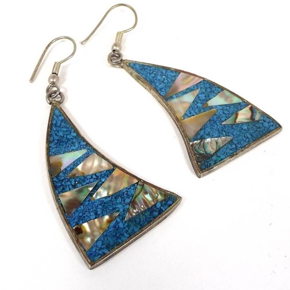 Front view of the Mexican Alpaca earrings. Earrings are triangle shaped with a curve. They are silver in color with aqua blue resin and tiny inlaid stone chips. There small triangle pieces of pearly multi color inlaid abalone shell that point inward from each side of the earring. They have fish hook style earwires at the top.