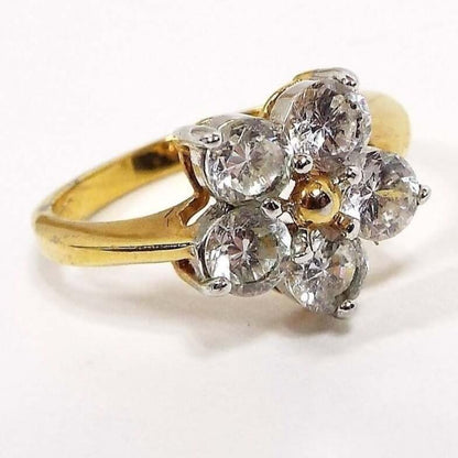 Angled front and side view of the retro vintage Edco rhinestone cocktail ring. The setting and band are gold tone in color. The ring has a flower design with five round clear rhinestones around a small gold tone ball. All stones are prong set. The sides of the band have an indented line at the top part of the ring. 