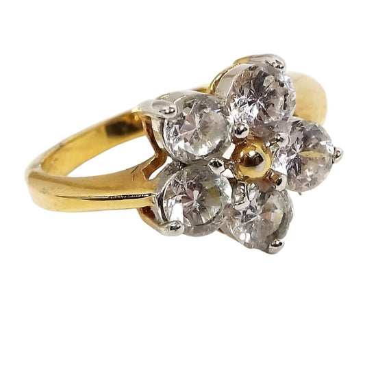Angled front and side view of the retro vintage Edco rhinestone cocktail ring. The setting and band are gold tone in color. The ring has a flower design with five round clear rhinestones around a small gold tone ball. All stones are prong set. The sides of the band have an indented line at the top part of the ring. 
