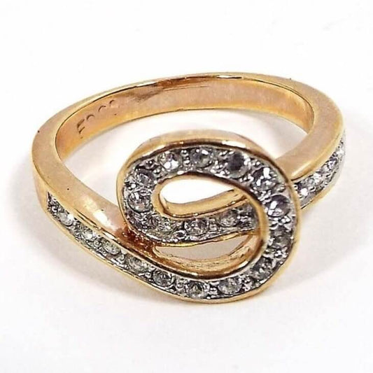 Angled front and side view of the retro vintage Edco rhinestone ring. The band and setting is gold tone in color. The top part of the band curves around to form a loose knot shape on the top. The knot and sides have a single row of pavé set round clear rhinestones. 