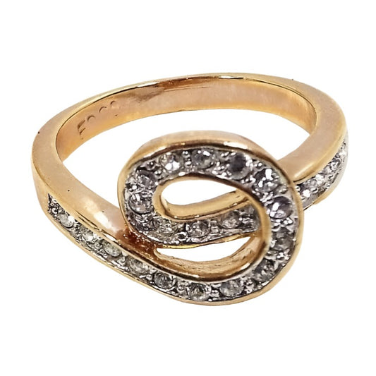Angled front and side view of the retro vintage Edco rhinestone ring. The band and setting is gold tone in color. The top part of the band curves around to form a loose knot shape on the top. The knot and sides have a single row of pavé set round clear rhinestones. 