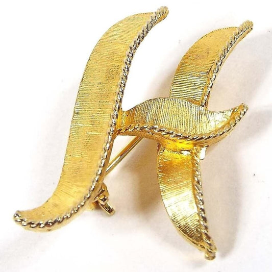 Front view of the Mid Century vintage letter brooch pin. The metal is matte brushed textured gold tone in color. It has a wavy block initial design of the letter H.