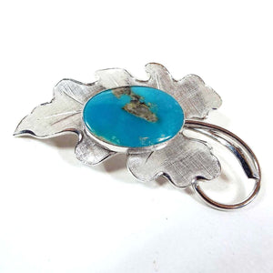 Front view of the Mid Century vintage Carl Art sterling leaf brooch pin. The leaf has a curvy triangle shaped pointed end design with matte brushed textured sterling. The stem is shiny sterling and is curled back towards the leaf. The middle has a large oval gemstone cab. The unidentified gemstone is turquoise blue in color with some gray rough stone area in the middle and towards one edge. The cab is bezel set. 