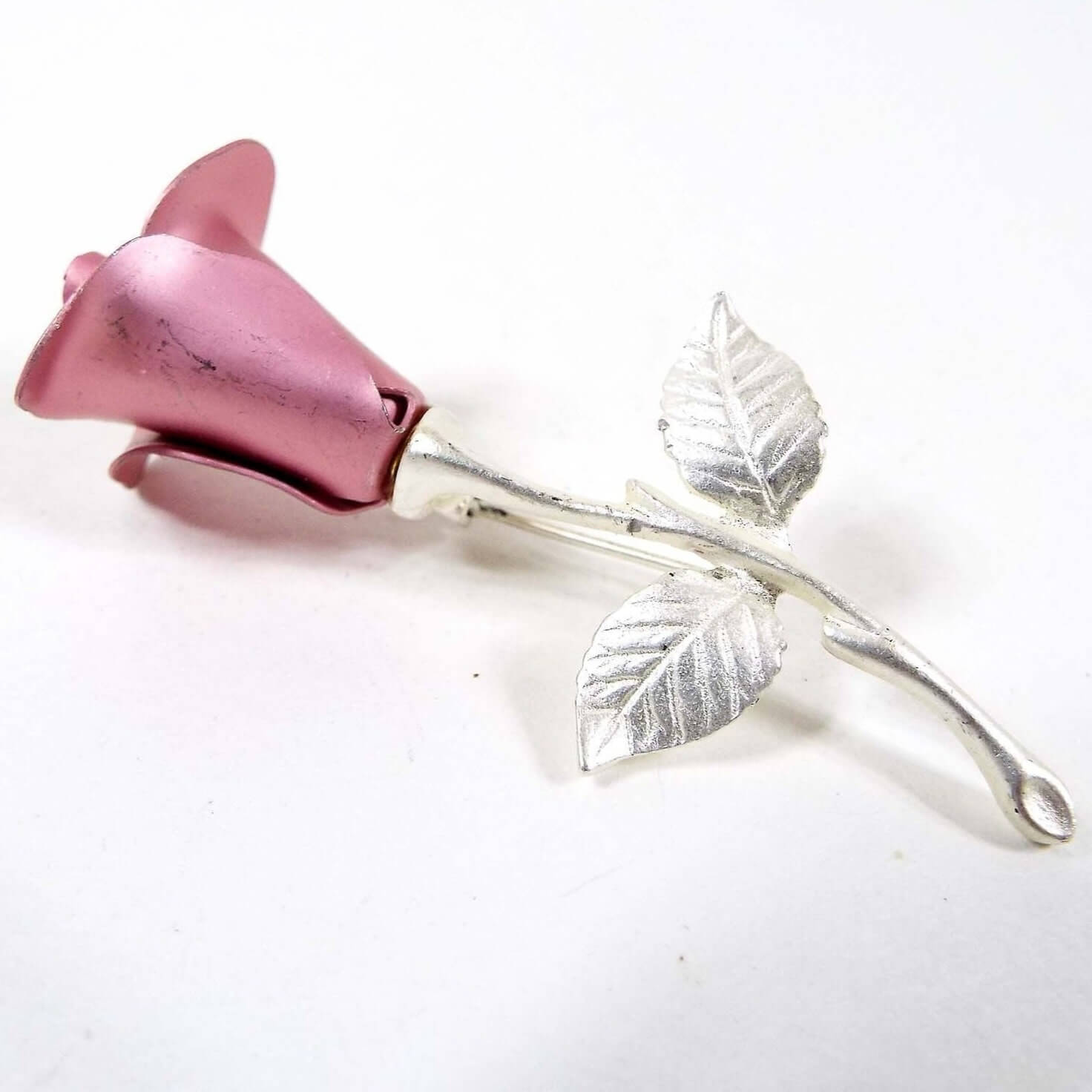 Front view of the retro 1990's Avon vintage rose flower brooch. It has a single rose design. The petals are pink painted aluminum. The stem, leaves, and pin are silver tone in color. 