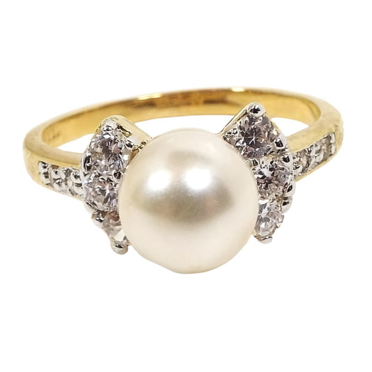 Angled front and side view of the retro vintage Edco cocktail ring. There is a larger sized round off white faux pearl made of plastic surrounded by a curved row of three round cubic zirconia stones on each side. The band has two small round CZ stones at the top on each side. The metal is gold tone in color. All of the CZ stones are prong set.