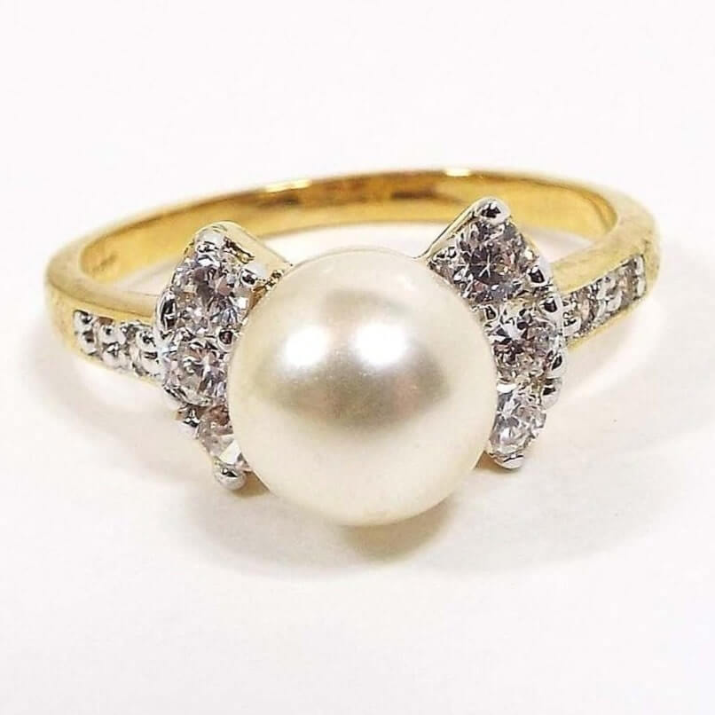 Angled front and side view of the retro vintage Edco cocktail ring. There is a larger sized round off white faux pearl made of plastic surrounded by a curved row of three round cubic zirconia stones on each side. The band has two small round CZ stones at the top on each side. The metal is gold tone in color. All of the CZ stones are prong set.