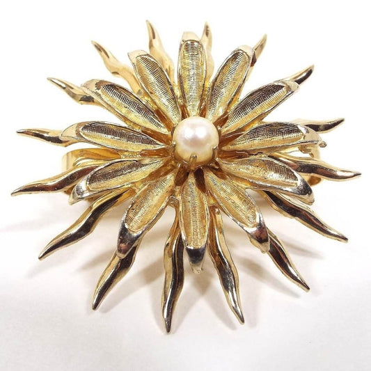 Front view of the retro vintage cultured pearl scarf clip. It has a floral design with the cultured pearl in the middle. It has two sets of petals for the flower. The first set of petals are pointed downward at the ends and have a matte brushed gold tone color. In between those are thinner spiky curved petals that have a shiny gold tone color. 
